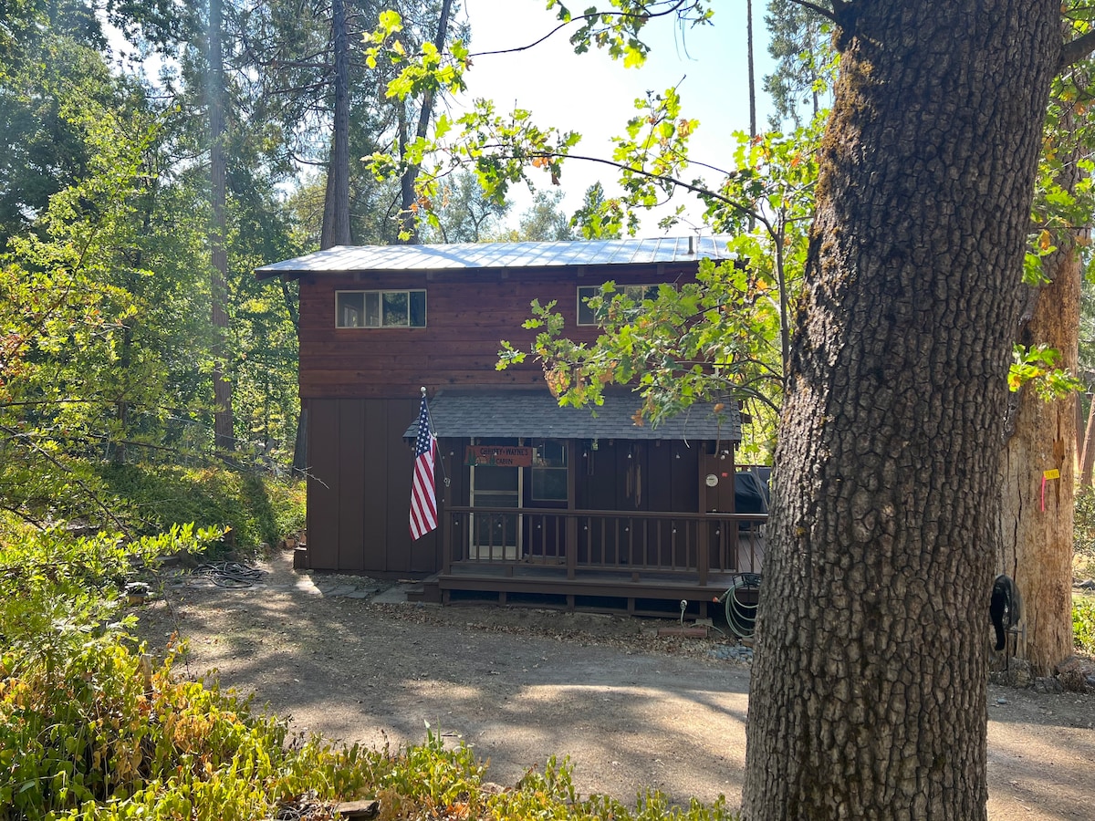 The exterior of a two-level cabin is framed by mature trees, showcasing a rustic wooden facade with an American flag displayed on the porch. A gravel path leads to the entrance, surrounded by lush greenery typical of the Sierra National Forest.