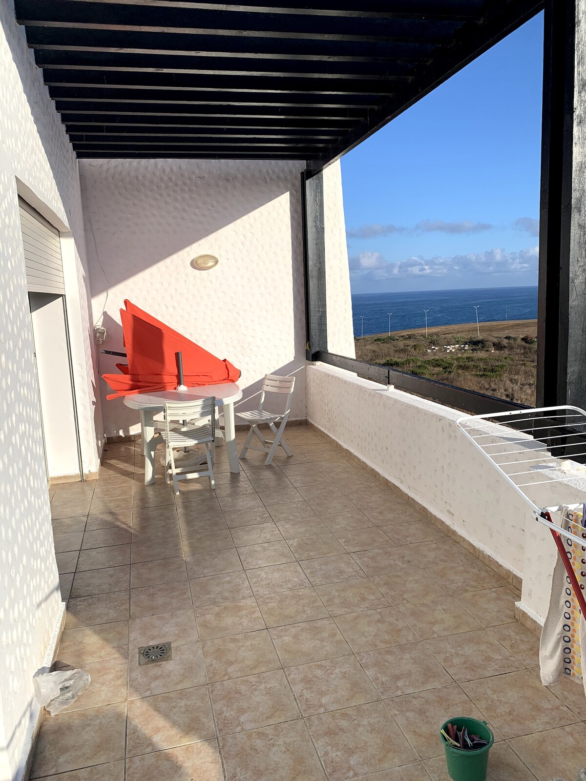 A sunlit balcony features a round table with four white chairs and is complemented by a vibrant orange tablecloth. The tiled floor extends across the space, while large windows frame a view of the distant ocean and sky.