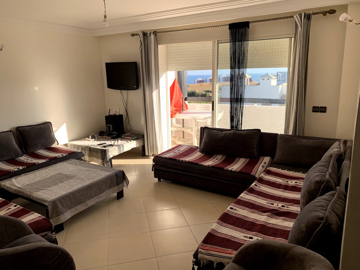 A spacious living area is furnished with multiple comfortable seating arrangements, including large sectional sofas. Large windows provide natural light and a view of the surroundings. A television is mounted on the wall, and a coffee table is centered in the room.