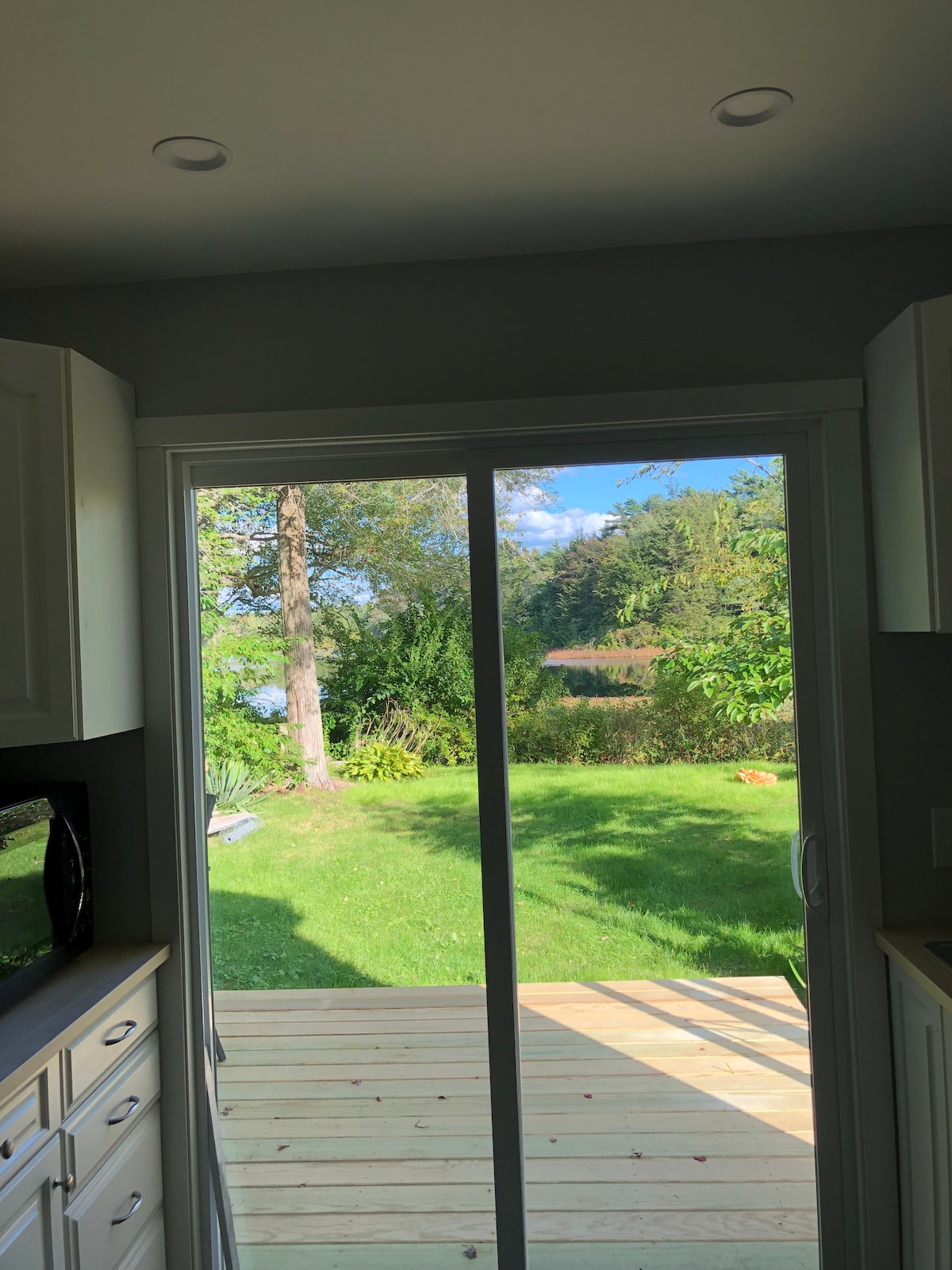 A sliding glass door frames a view of the lush green yard, featuring a large tree and a glimpse of the lake beyond. Sunlight filters through the foliage, illuminating the space while creating a serene outdoor atmosphere.