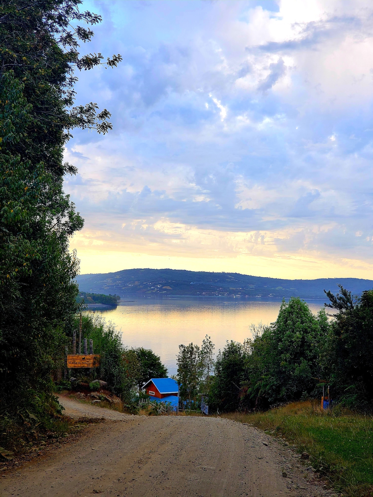 A dirt road descends gently towards a serene body of water, reflecting the soft colors of the sky. Lush greenery lines both sides of the path, while a house with a blue roof is visible at the shoreline, framed by trees and hills in the distance.