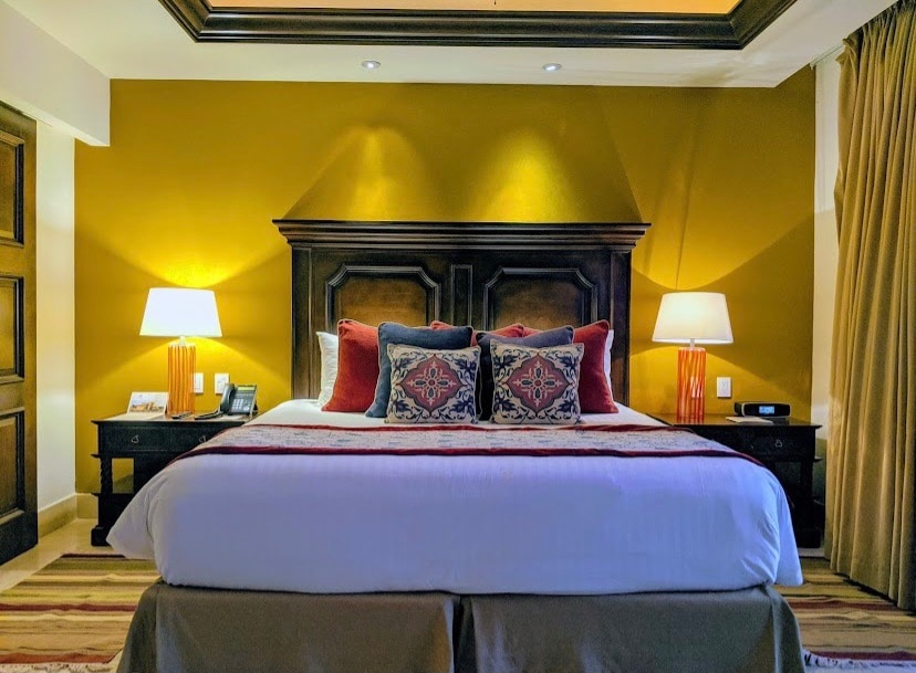 An elegantly furnished bedroom features a spacious bed adorned with decorative pillows, set against a warm yellow wall. Two bedside lamps provide soft lighting, complemented by dark wood nightstands. Rich curtains frame the large window, adding a touch of sophistication to the design.
