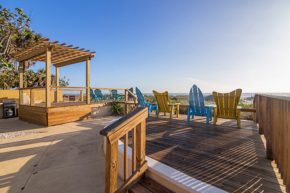 An outdoor patio area features a wooden deck with a shaded pergola. Colorful adirondack chairs and tables are arranged for relaxing, with views of the ocean in the background. Lush greenery adds a touch of nature to this inviting space.