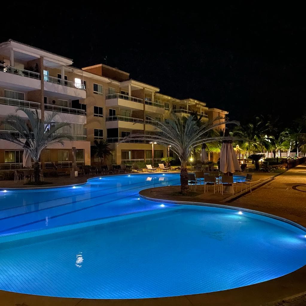 The illuminated pool area features a mix of vibrant blue waters surrounded by palm trees and lounge chairs. The three-story building stands in the background, showcasing its balconies, while warm lighting enhances the inviting atmosphere of the outdoor space.