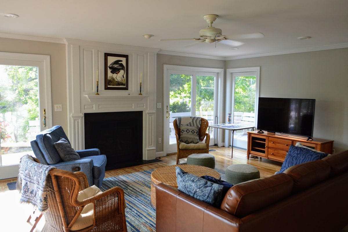 A cozy living room is highlighted by a fireplace and large windows that invite natural light. Comfortable seating includes a leather sofa, woven armchair, and a blue chair. A television is positioned opposite the sofa, and a patterned rug adds warmth to the space.