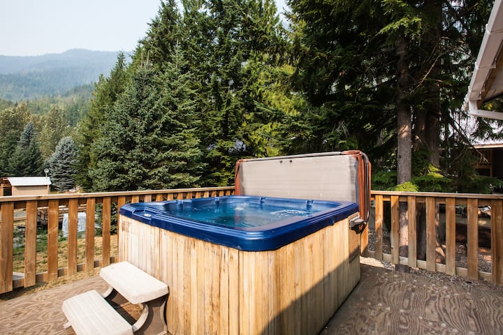 A large outdoor hot tub is surrounded by a wooden deck, positioned among tall evergreen trees. The tub's cover is lifted to reveal clear water, inviting relaxation in a natural setting. A simple bench is placed nearby for convenience.