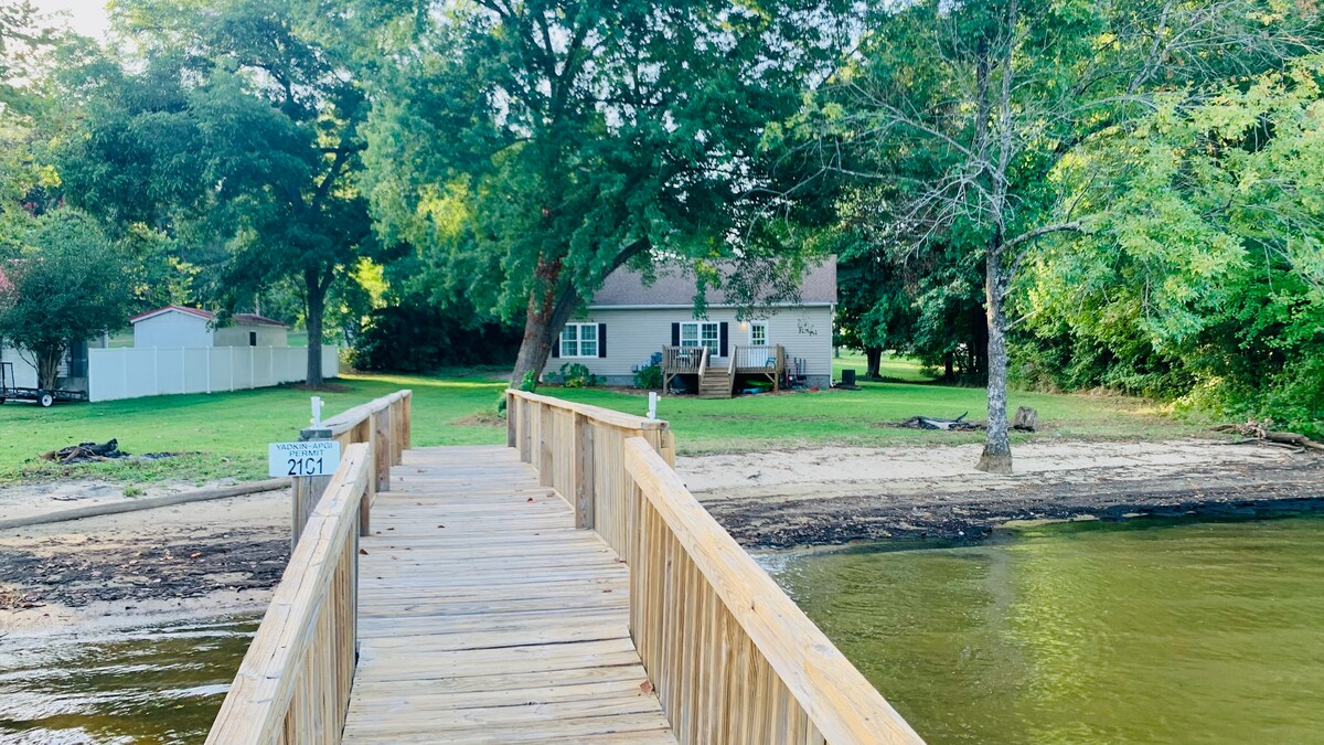 A wooden dock leads from the serene water to a charming single-story home surrounded by lush greenery. The well-maintained lawn extends to a sandy beach area, offering a tranquil outdoor space. Trees provide natural shade, contributing to the peaceful atmosphere.