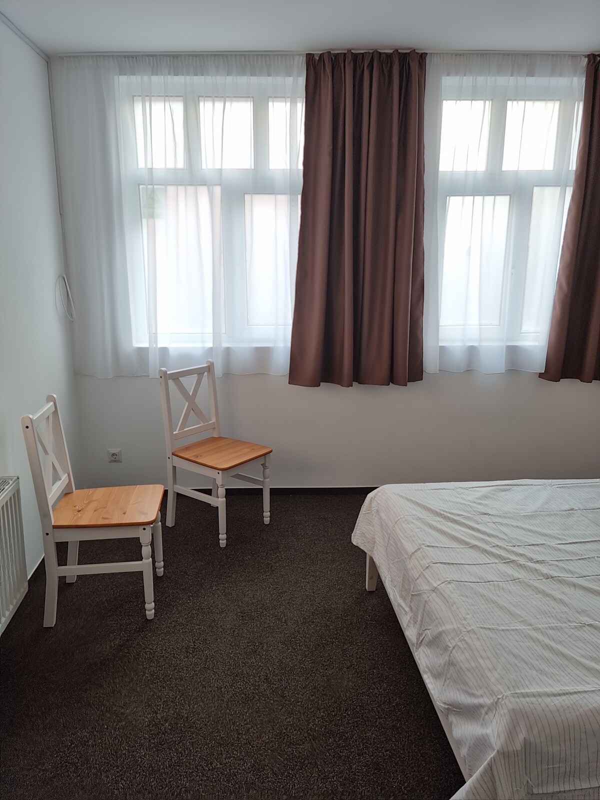 A bedroom features a spacious bed against a wall with three large windows, allowing natural light to fill the room. Two wooden chairs with simple design are positioned nearby, complemented by light curtains that softly diffuse incoming sunlight.