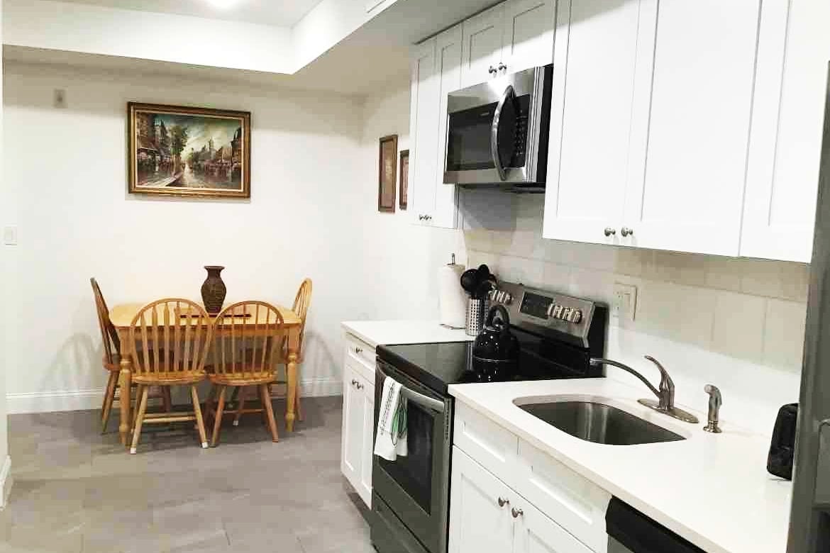 The kitchen area features white cabinetry and stainless steel appliances, including a microwave and oven. A dining table with wooden chairs is positioned adjacent to a decorative wall art piece. A sleek sink and countertop provide ample preparation space, illuminated by overhead lighting.