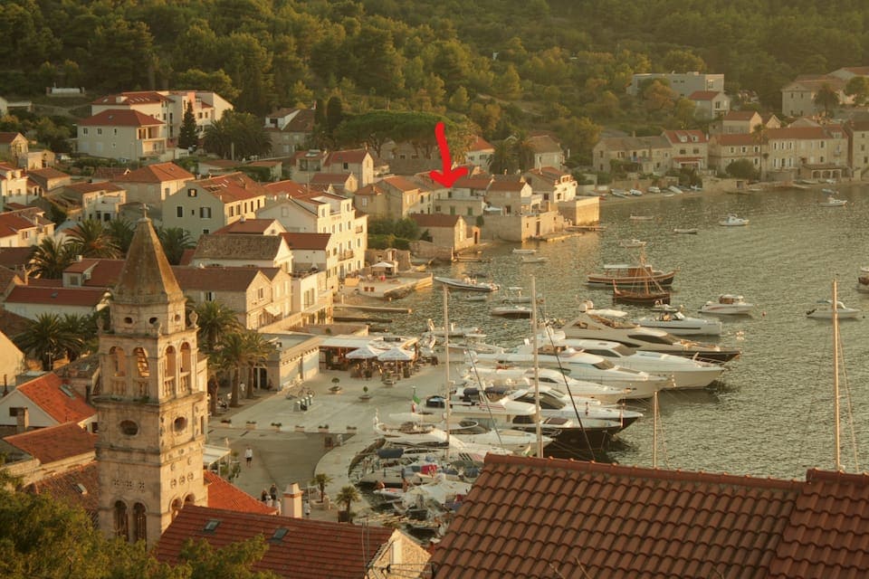 A panoramic view of the waterfront area showcases several boats anchored in the harbor. Nearby, charming stone houses are nestled among trees, with a prominent historic building featuring a tall clock tower in the foreground.