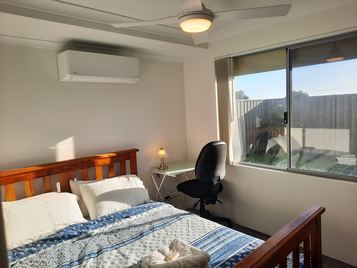 A well-lit bedroom features a wooden bed with soft linens and a decorative blanket. A desk and chair provide a workspace near a large window that offers views of the outdoor area, complemented by a ceiling fan and air conditioning unit.