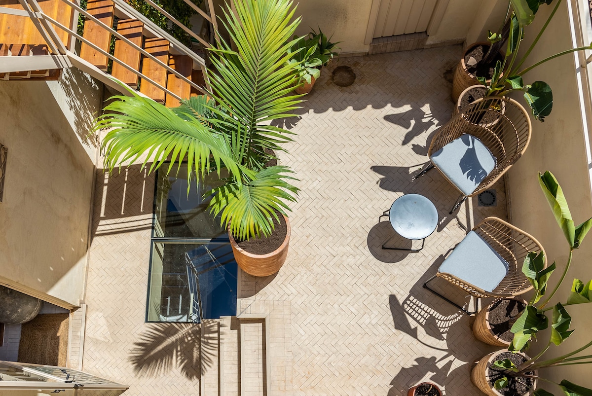 An outdoor terrace is visible, featuring a glass table surrounded by two chairs. Potted plants add greenery, and sunlight creates playful shadows on the herringbone-patterned floor. A staircase can be seen leading to the upper levels, enhancing the open and airy feel of the space.