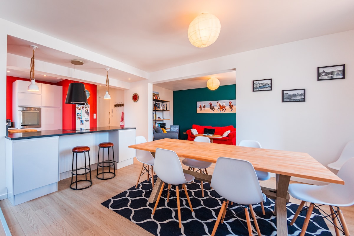 A modern dining area features a large wooden table surrounded by white chairs. A stylish kitchen bar with three stools complements the space. Light fixtures hang overhead, while a cozy living area with red accents can be glimpsed in the background.