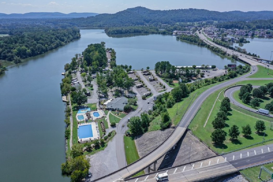 An aerial view captures a scenic landscape with the Coosa River winding gracefully alongside a recreational area. Multiple pools are visible, surrounded by shaded trees and neatly arranged spaces, while hills rise gently in the background.