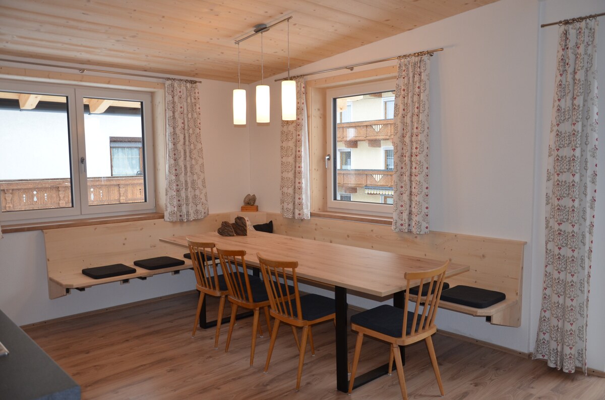 A bright dining area is furnished with a long wooden table and six wooden chairs, positioned against a wall with large windows. Soft curtains frame the windows, allowing natural light to illuminate the space, accentuated by three hanging light fixtures.