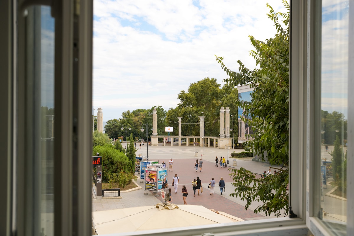 A view of a bustling public square is captured from an open window. Pedestrians can be seen strolling along the pathway, surrounded by trees and greenery. The architectural elements of the square are visible, contributing to the lively atmosphere.