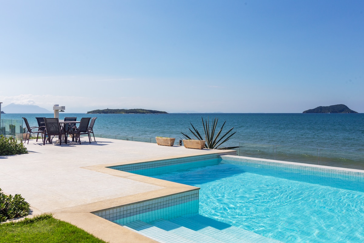 A tranquil outdoor space is showcased, featuring a clear swimming pool with a deck that overlooks the ocean. A dining table and chairs are positioned on the patio, inviting relaxation with a serene view of distant islands and a bright sky.