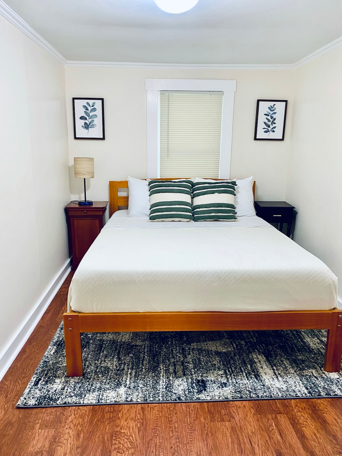 A cozy bedroom features a wooden bed with a light-colored bedspread. Two decorative pillows with stripes add detail to the bed. A nightstand and lamp provide soft lighting. Framed botanical prints hang on the walls, and a small window allows natural light to fill the space.