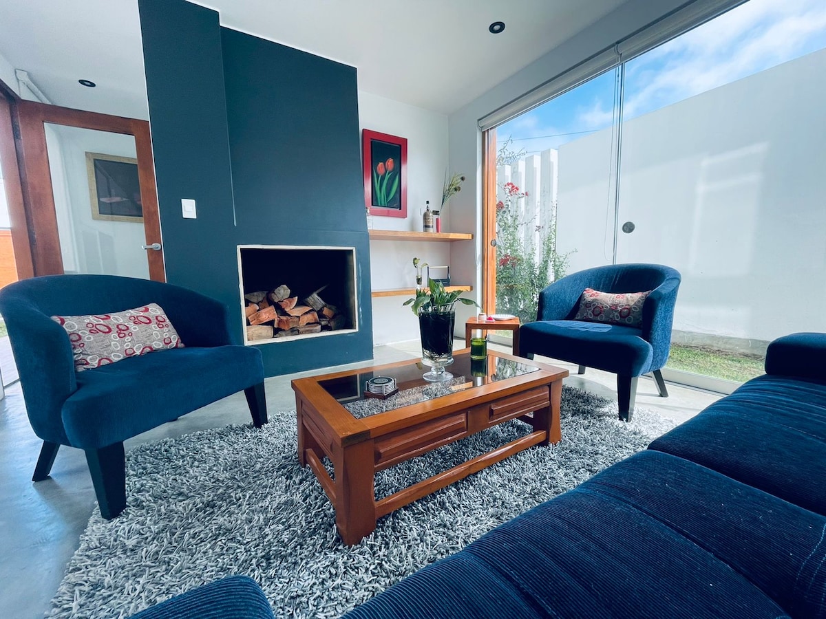 A comfortable living area features two blue armchairs and a dark sofa, arranged around a wooden coffee table. A cozy fireplace is situated against a dark accent wall. Natural light enters through large sliding glass doors, highlighting a small garden visible outside.