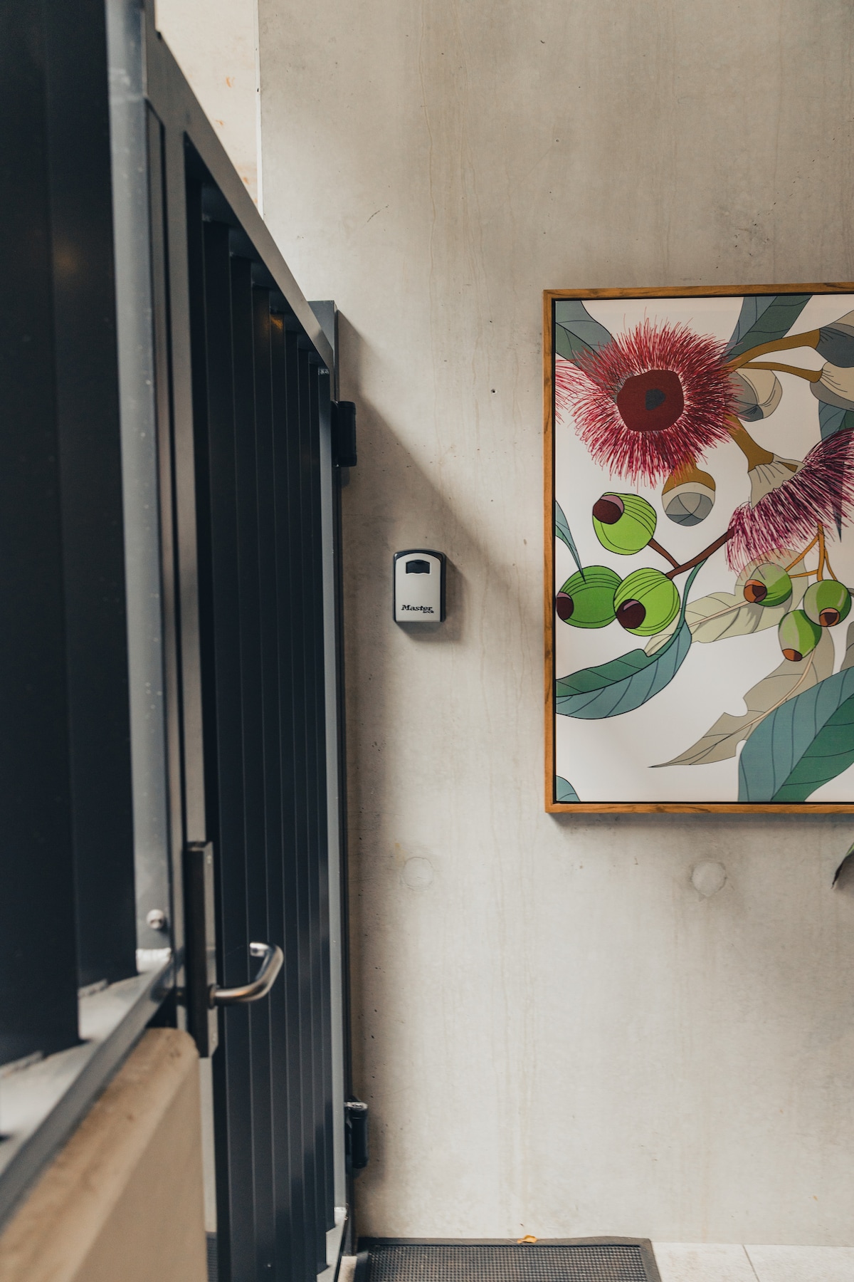 A modern entryway features a sleek black door alongside a colorful artwork depicting floral and botanical elements. A contemporary light fixture is mounted on the wall, complementing the concrete surfaces and creating a minimalist aesthetic.