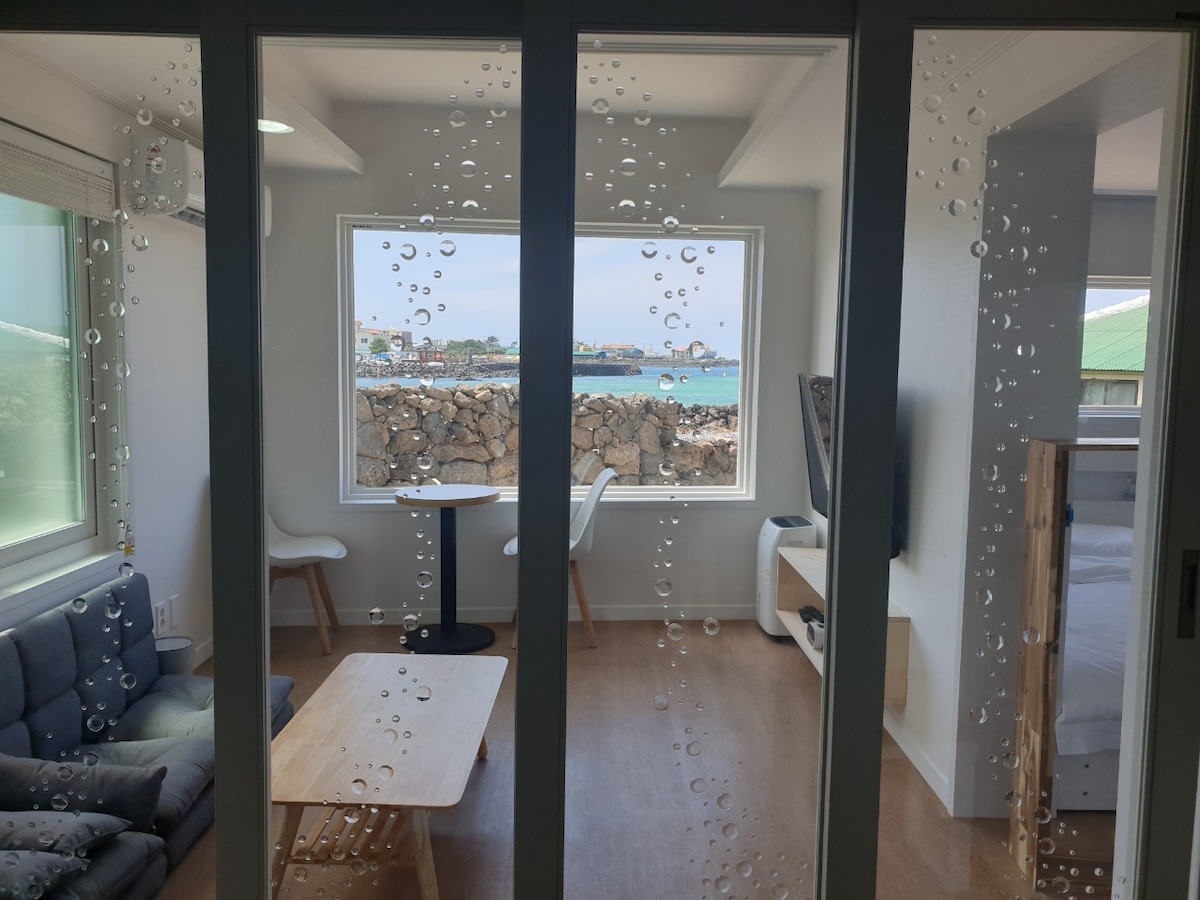 A bright living space is visible through glass doors adorned with water droplets. A cozy seating area features a gray couch facing a small round table and two white chairs. A view of the sea and distant buildings can be seen through the large window.