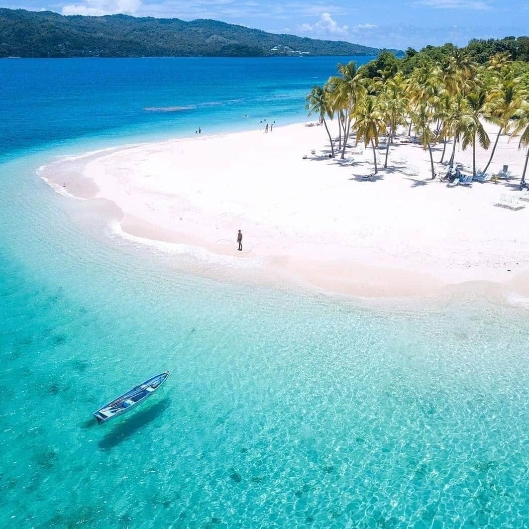 A serene beach scene is captured, featuring soft white sands bordered by clear, turquoise waters. A small boat floats gently near the shoreline, while a few people enjoy the sun and palm trees provide shade in the background. The landscape is set against a backdrop of lush green hills.