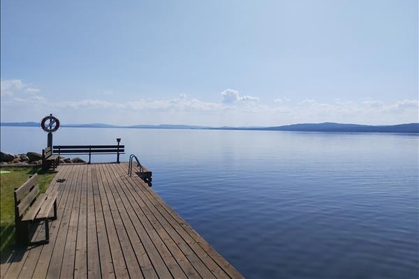 A wooden dock extends into calm, reflective water, surrounded by a clear blue sky. Nearby, a lifebuoy is mounted on a post, while benches provide seating along the shore. The tranquil setting emphasizes the natural beauty of the lakeside.