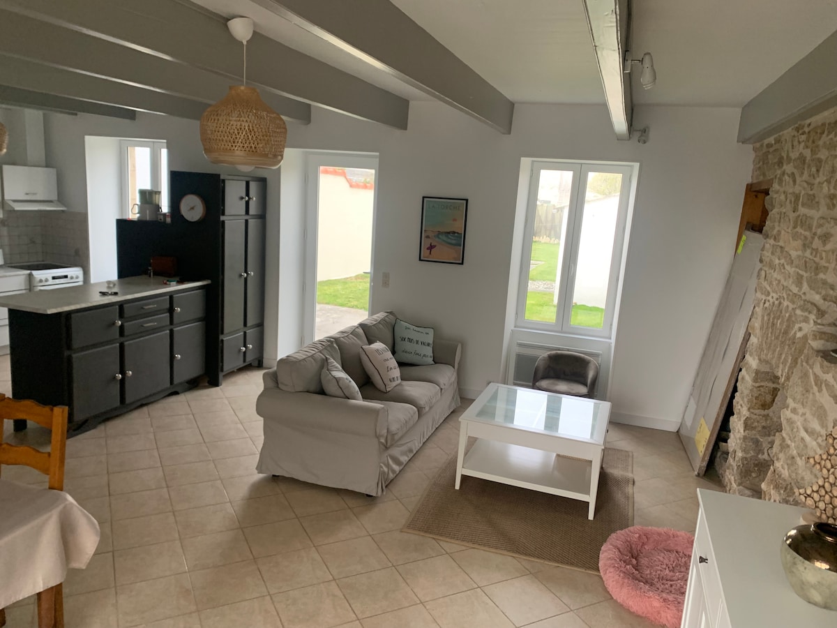 A living area showcases a light-colored sofa and a glass coffee table on a soft rug. Natural light streams through two windows, illuminating the space. The adjoining kitchen area features dark cabinetry and modern appliances, while stone walls add character to the environment.