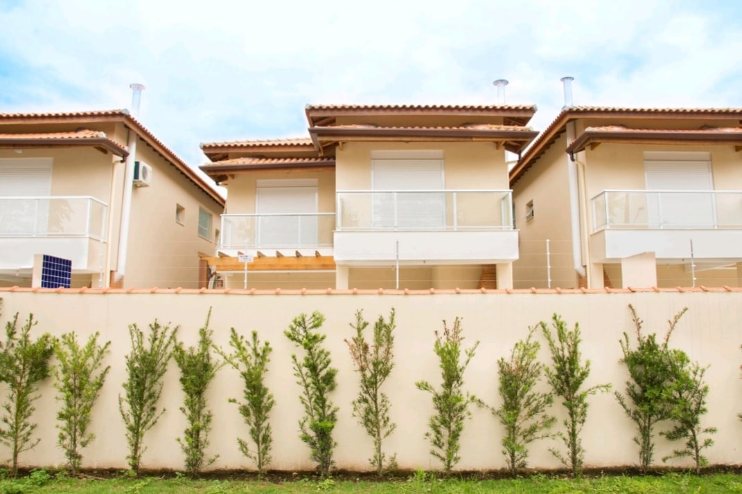 A row of three modern houses is displayed against a clear blue sky, showcasing balconies with glass railings. A neatly maintained hedge runs along the property's boundary, complementing the clean lines of the buildings.