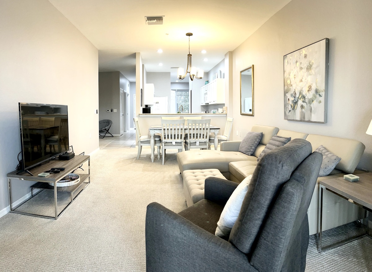 A modern living area features a plush sectional sofa and a cozy armchair. A sleek TV is placed on a metal stand, while a dining table with white chairs is positioned in the background. Light from overhead fixtures brightens the space, complemented by neutral wall tones.