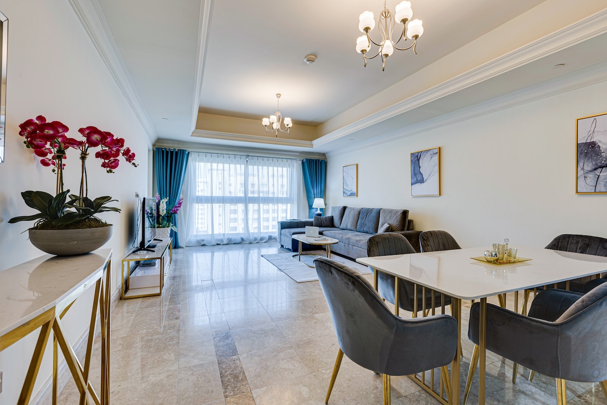 A spacious living area features a large sofa and elegant dining table. Natural light filters through floor-to-ceiling windows adorned with blue drapes. Decorative plants and artwork add a touch of refinement, while polished marble flooring enhances the overall ambiance.