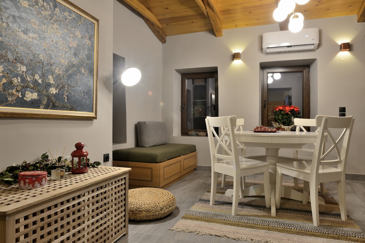 A dining area is presented with a round table surrounded by four white chairs. A cozy seating nook is positioned nearby, complemented by a decorative piece on the wall. Soft lighting enhances the inviting atmosphere, while a rug adds texture to the floor.