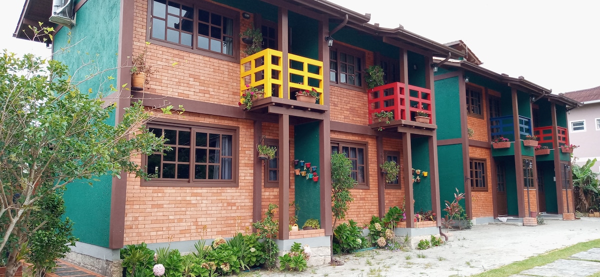 A row of charming brick apartments is displayed, featuring colorful balconies in red, yellow, and blue. Lush greenery and potted plants adorn the building's façade, creating a welcoming outdoor environment. Large windows provide ample natural light, enhancing the inviting character of the space.