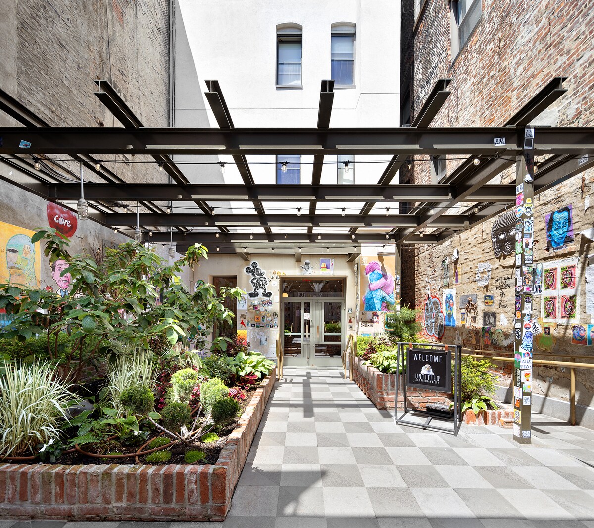 An inviting courtyard is framed by a metal canopy, featuring potted plants and artwork displayed on red brick walls. A checkerboard pattern decorates the floor, leading towards an entrance with a welcoming sign. Colorful illustrations add character to the surrounding space.