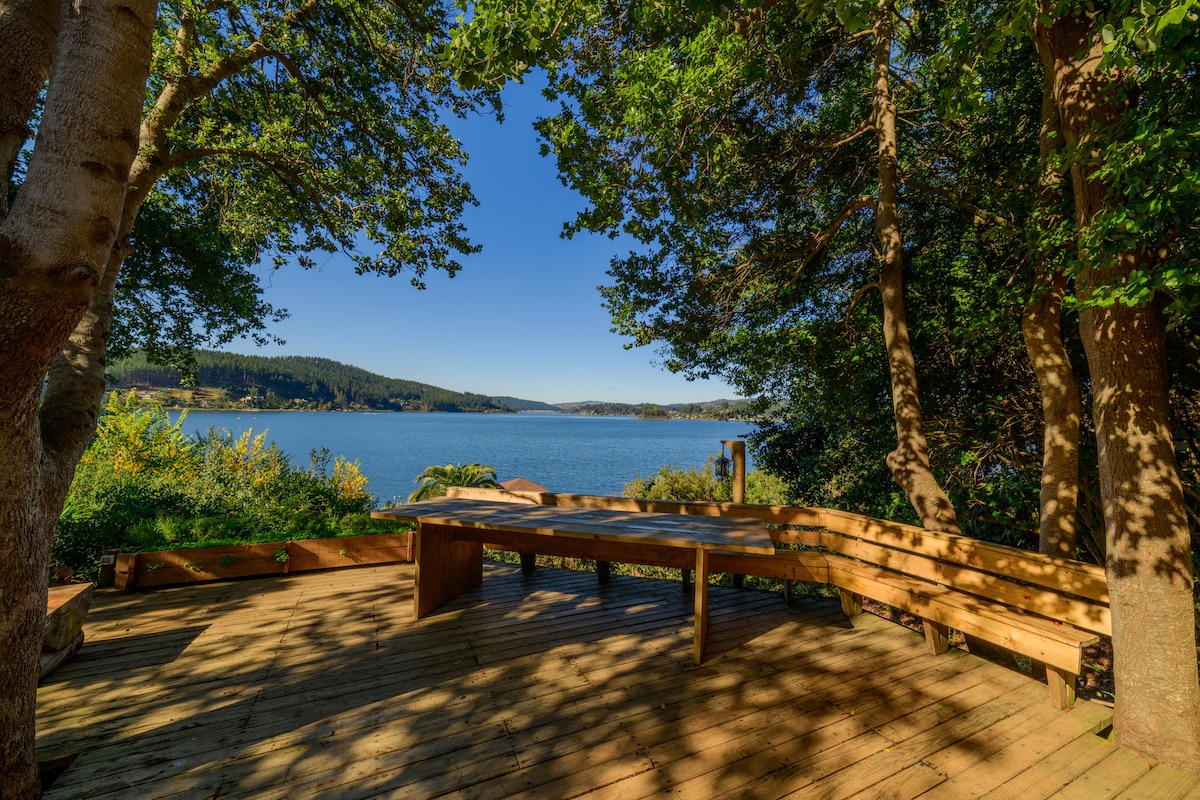 A spacious wooden terrace is surrounded by trees, offering a direct view of a serene lake. The surface is complemented by built-in seating, providing a comfortable area to relax and enjoy nature. The clear sky enhances the peaceful ambiance of the outdoor setting.