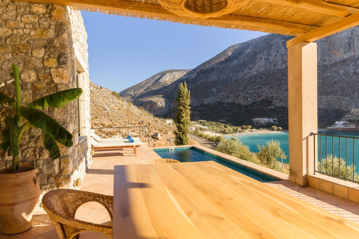 An outdoor dining area is set under a wooden pergola, featuring a large wooden table. In the background, a private pool and stunning sea views are framed by surrounding mountains and olive trees.