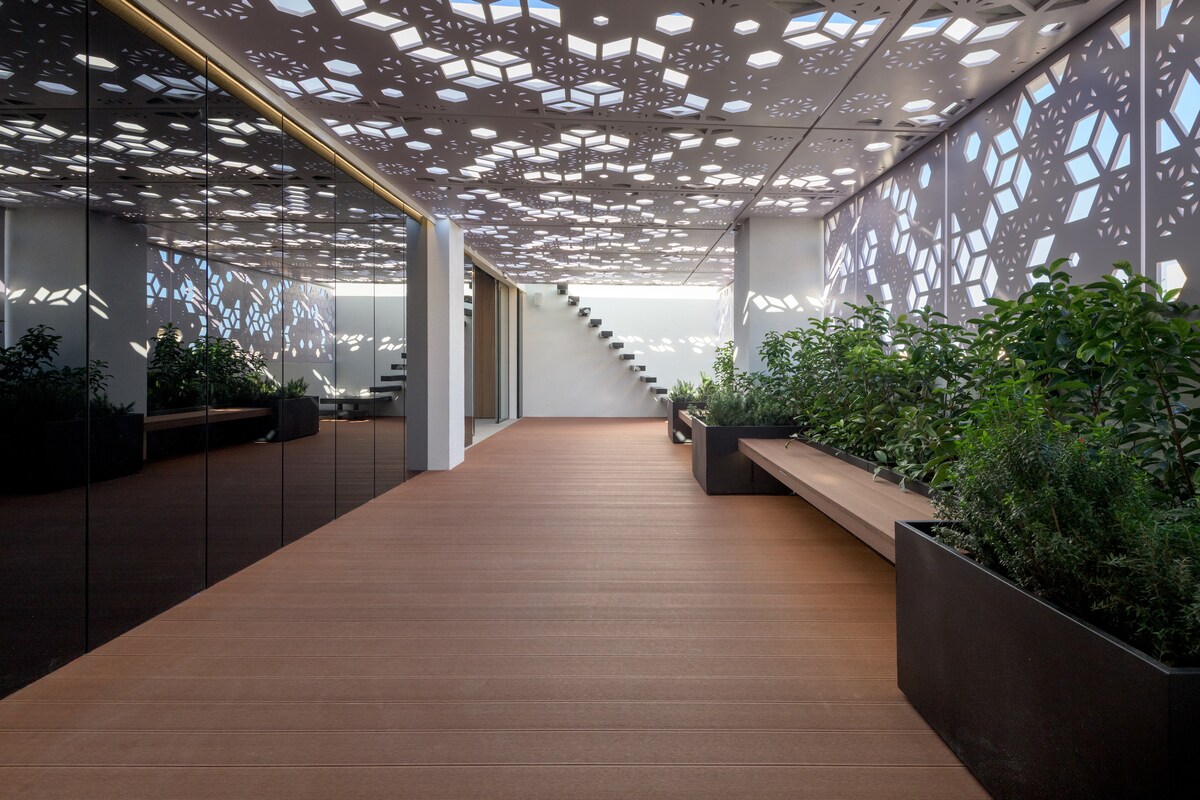 A modern corridor is illuminated by intricate ceiling designs that filter natural light. Smooth wooden flooring extends through the space, complemented by reflective glass walls. Lush greenery is found in built-in planters along one side, creating a serene environment that invites peaceful transition.