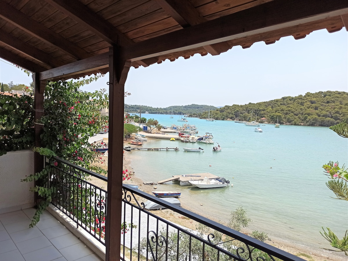 A view of a tranquil bay is showcased from a spacious balcony. Various boats are anchored in the water, with lush greenery lining the shore. The sturdy wooden beams and railing add a rustic charm to the setting, capturing the essence of a peaceful getaway.