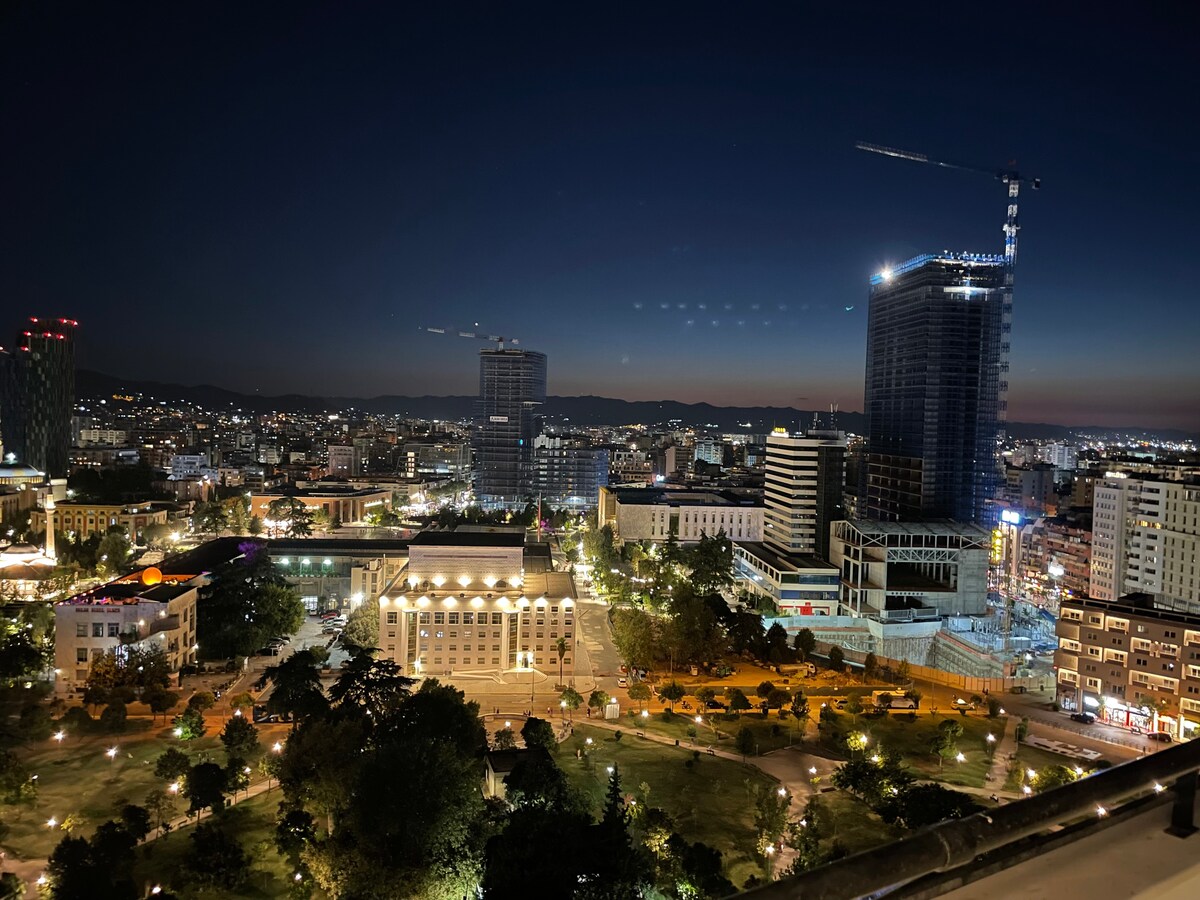 A cityscape is captured at night, featuring illuminated buildings and busy streets. The scene highlights a prominent park at the center, surrounded by modern high-rises and soft lighting that outlines walkways. The distant mountains form a backdrop against the night sky.