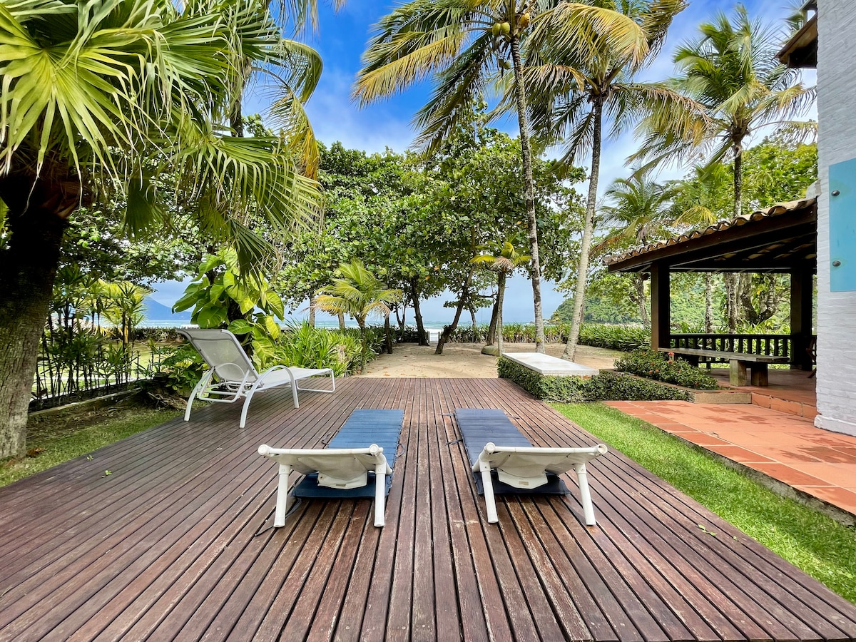 A wooden deck is bordered by lush greenery and palm trees, featuring two sun loungers positioned for relaxation. The view opens up towards a tranquil garden area with distant glimpses of the beach and ocean beyond, creating a serene outdoor setting.