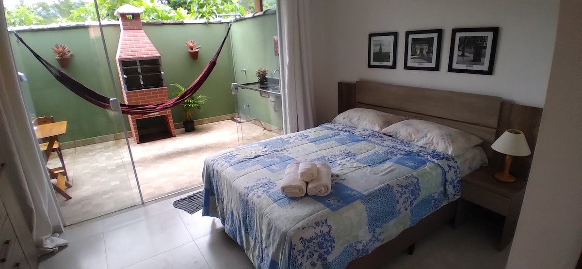 A spacious loft features a comfortable bed adorned with a patterned blanket, alongside a bedside lamp and decorative artwork on the wall. Glass doors open to a private patio area with a hammock and a barbecue grill, creating an inviting outdoor space.