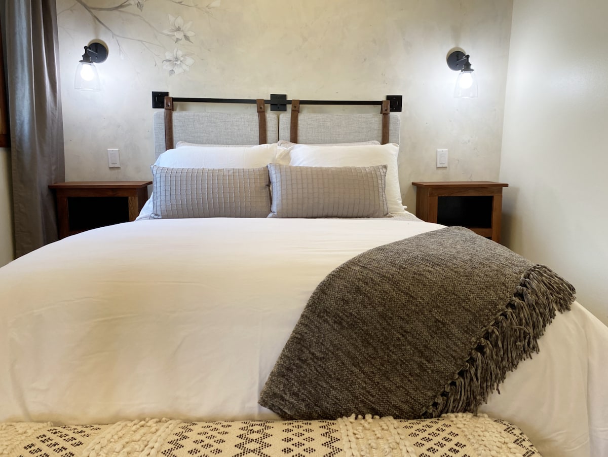 A spacious bed is featured with crisp white linens and decorative throw pillows. Natural light filters through adjacent curtains, illuminating the room. Simple bedside tables provide a functional aspect, and stylish wall sconces add subtle lighting, enhancing the overall comfort and practicality of the space.