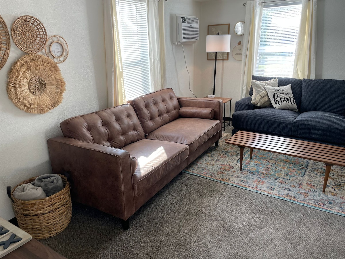 A cozy living area is presented, featuring a brown leather couch and a dark blue fabric sofa. A wooden coffee table sits in front, with an area rug adding texture to the floor. Natural light filters through the windows, illuminating woven wall decor.