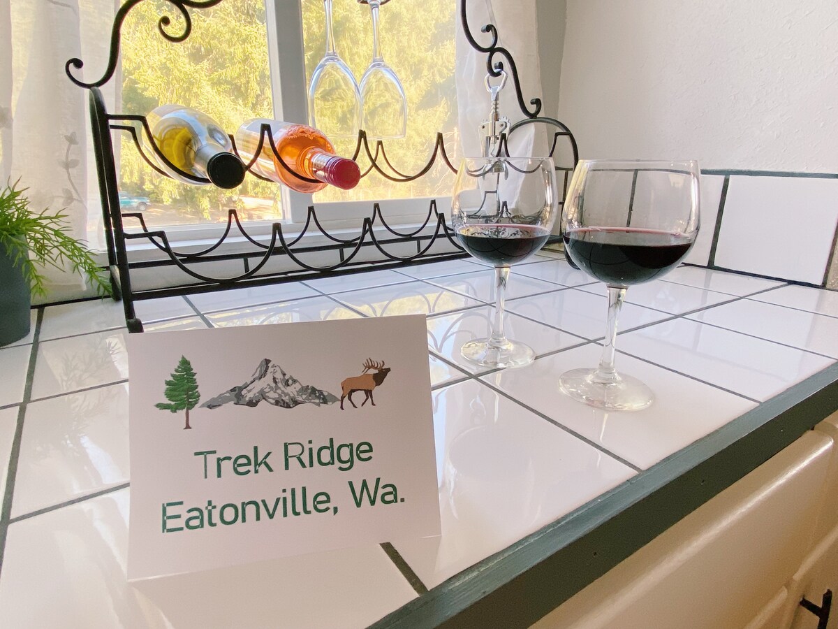 A kitchenette counter is presented with two glasses of red wine alongside a sign displaying 'Trek Ridge Eatonville, Wa.' A wine rack holds several bottles in the background, and natural light filters through a nearby window.