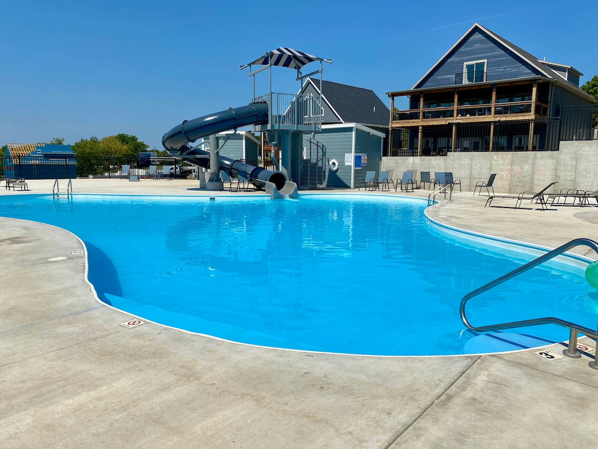 The outdoor pool area features a large, clear blue swimming pool with a gradual entry. A waterslide is visible nearby, along with several lounge chairs positioned around the pool deck. The backdrop includes a multistory building, hinting at guests' accommodations.