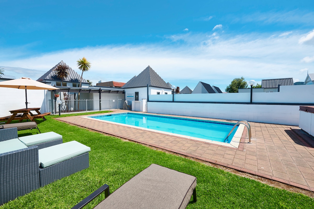 An outdoor saltwater pool is framed by a spacious area of manicured grass. Comfortable lounge chairs are positioned nearby, alongside a shaded seating area under an umbrella. The surrounding landscape includes modern structures and clear blue skies, inviting relaxation and leisure.