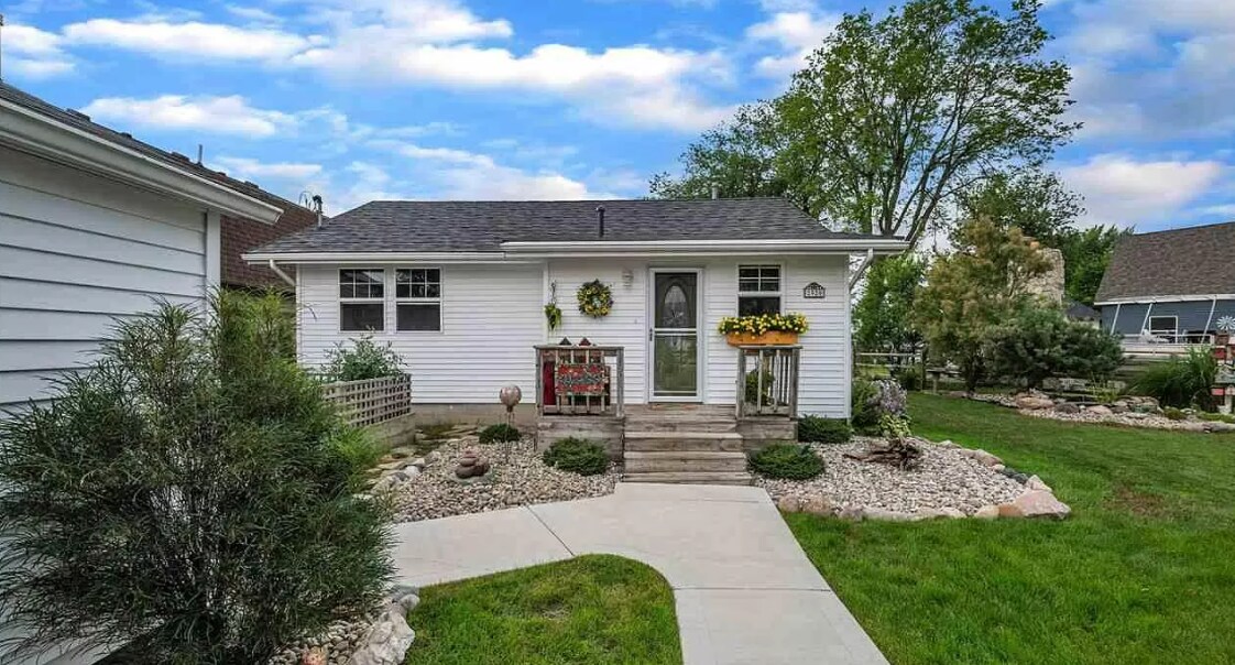 A welcoming exterior features a charming white house surrounded by well-maintained landscaping. Flower boxes adorn the windows, and a stone pathway leads to the entrance, enhancing the inviting atmosphere. Lush green grass and decorative stones frame the home.
