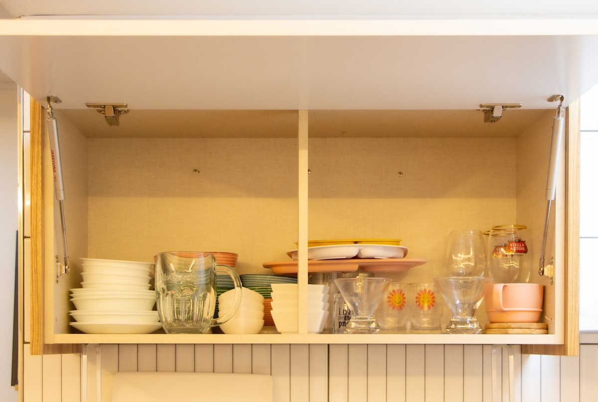A neatly organized kitchen cabinet displays a variety of dishware. White plates are stacked alongside colorful bowls and glasses. Various serving platters and decorative items are arranged, creating an accessible space for meal preparation and dining.