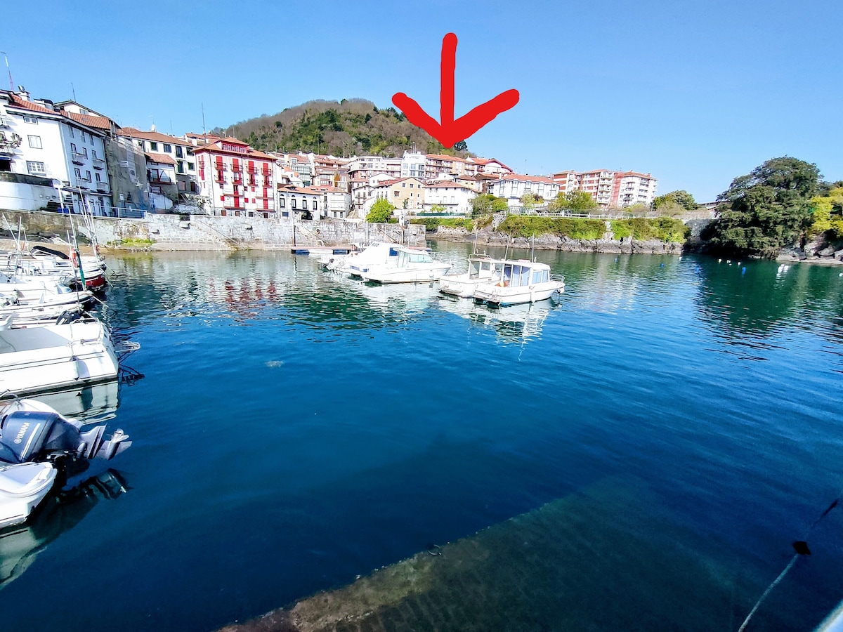 A serene waterfront view captures several boats anchored in calm waters, reflecting the surrounding landscape. In the distance, a charming village is nestled on a hillside, showcasing traditional architecture amidst greenery.