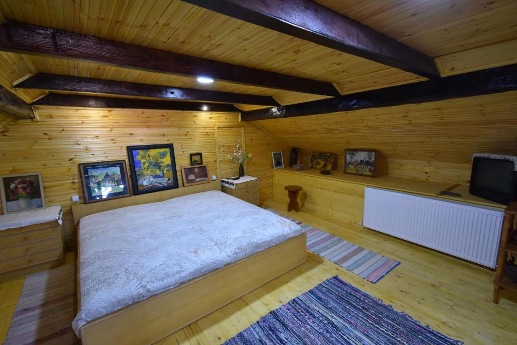 A spacious bedroom features wooden walls and beams, providing a warm ambiance. A large bed with a light-colored quilt is centered on the wooden floor. Artwork is displayed on the walls, and a radiator is present beneath a window. Area rugs add texture to the space.